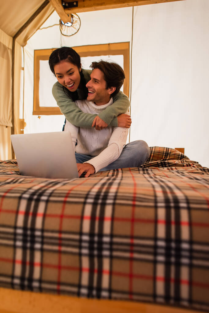 Alegre asiático mujer abrazando novio freelancer en cama en glamping casa  - Foto, Imagen