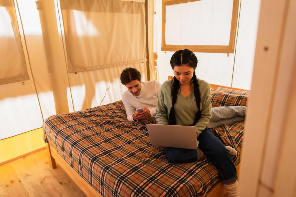 Asiático freelancer usando laptop cerca de novio con smartphone en cama en glamping house  - Foto, Imagen