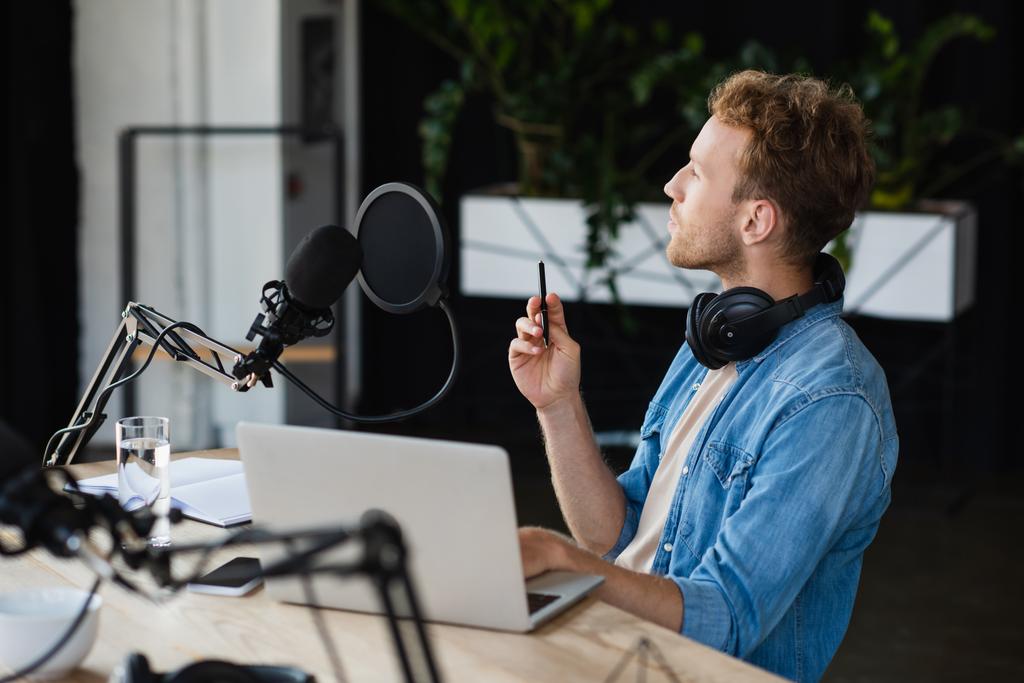 anfitrião de rádio pensativo em fones de ouvido segurando caneta perto de gadgets e microfone em estúdio  - Foto, Imagem