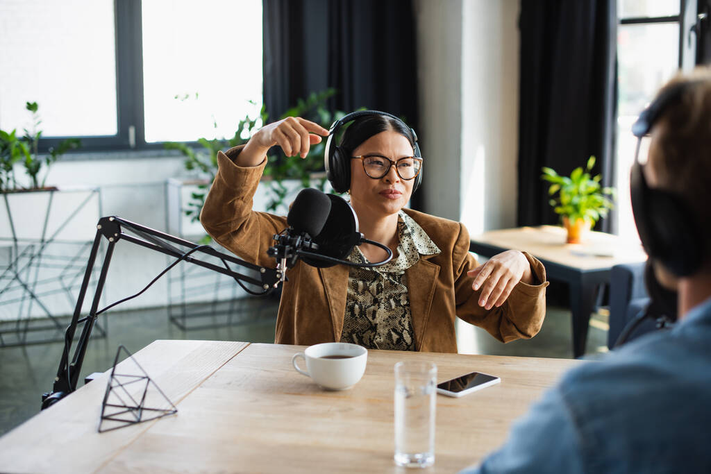 Asijské rádio hostitel v brýlích a sluchátka gestikulace v blízkosti rozmazané kolegy ve studiu - Fotografie, Obrázek