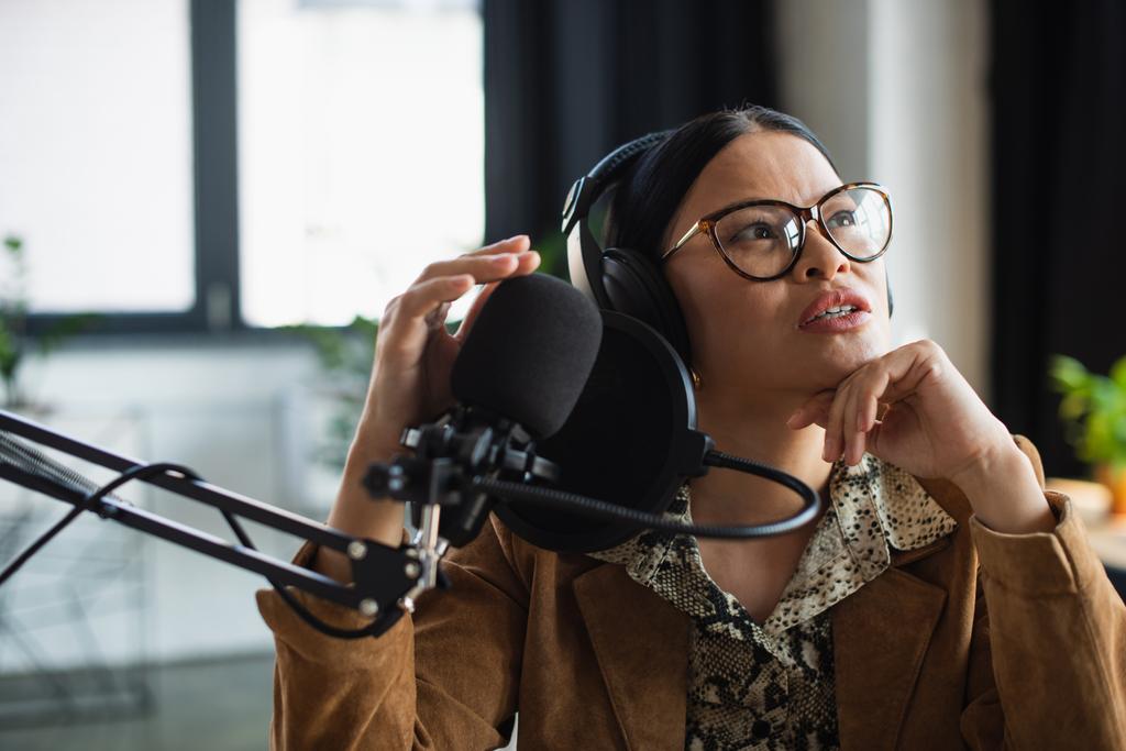 asiático radio host en gafas y auriculares mirando arriba mientras hablando en broadcast studio - Foto, Imagen