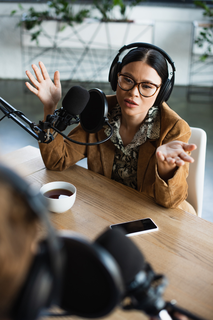alto ángulo vista de asiático radio host en gafas y auriculares gesto cerca de taza de café y borrosa colega durante podcast - Foto, imagen