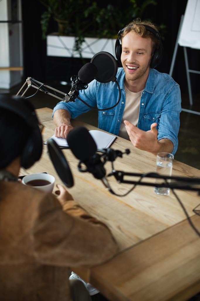 cheerful radio host in headphones looking at blurred colleague and gesturing during podcast - Photo, Image