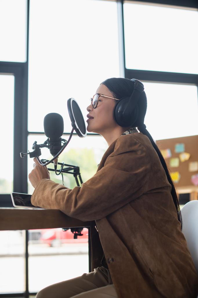 widok z boku azjatyckiego hosta radiowego w okularach i słuchawkach rozmowy podczas podejmowania podcast w studio - Zdjęcie, obraz