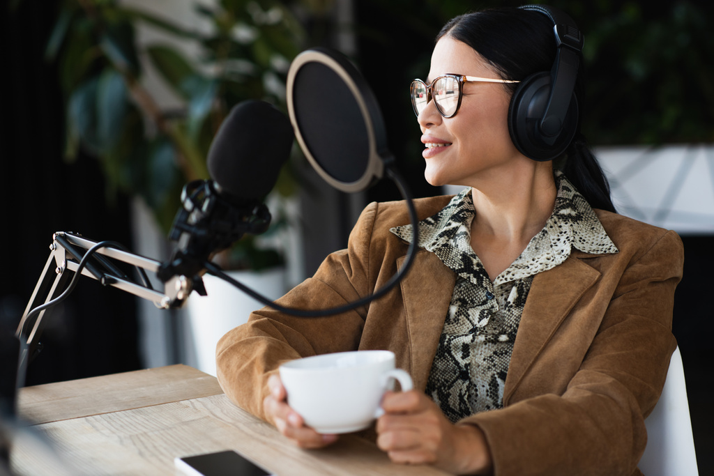 elégedett ázsiai rádió házigazda szemüvegben és fejhallgató kezében csésze kávét podcast alatt  - Fotó, kép