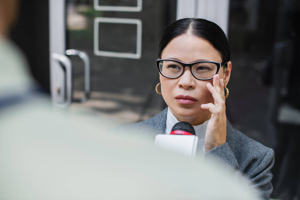 borrosa reportera con micrófono cerca pensativo asiático mujer de negocios ajuste gafas  - Foto, imagen