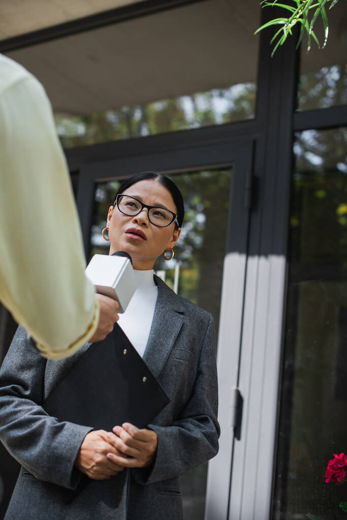 dziennikarka trzymająca mikrofon i przeprowadzająca wywiad z azjatycką bizneswoman w okularach - Zdjęcie, obraz
