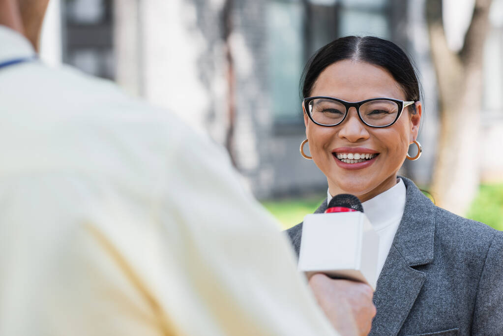 reporter floue debout avec microphone près positive asiatique femme d'affaires dans des lunettes  - Photo, image