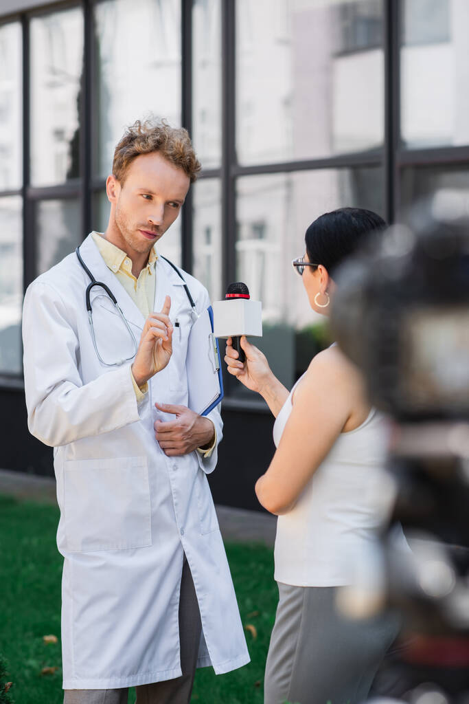 orvos fehér kabátban ujjal mutogatás és figyelmeztetés, miközben interjút ázsiai újságíró mikrofonnal  - Fotó, kép
