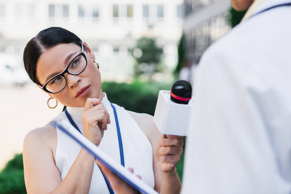 брюнетка азіатський журналіст тримає мікрофон і дивиться на розмитого лікаря в білому пальто
  - Фото, зображення