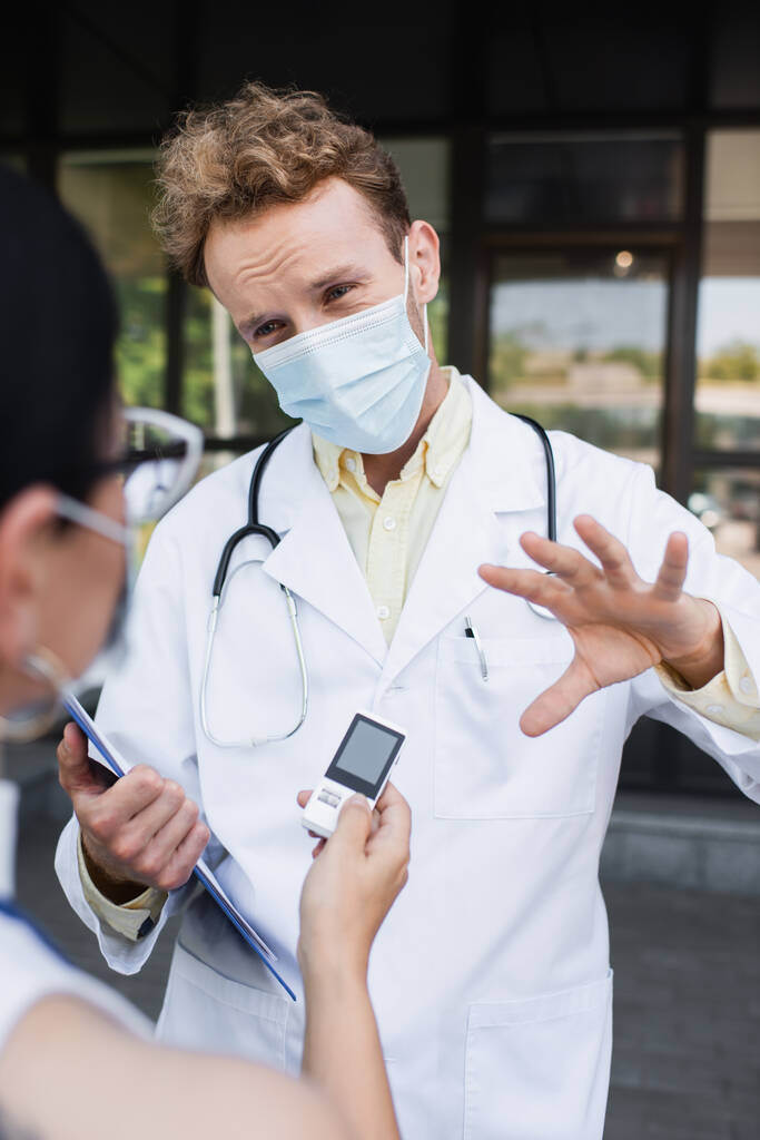 zamazany azjatycki reporter trzymając rejestrator głosu w pobliżu lekarza w masce medycznej i biały płaszcz gesting podczas wywiadu  - Zdjęcie, obraz