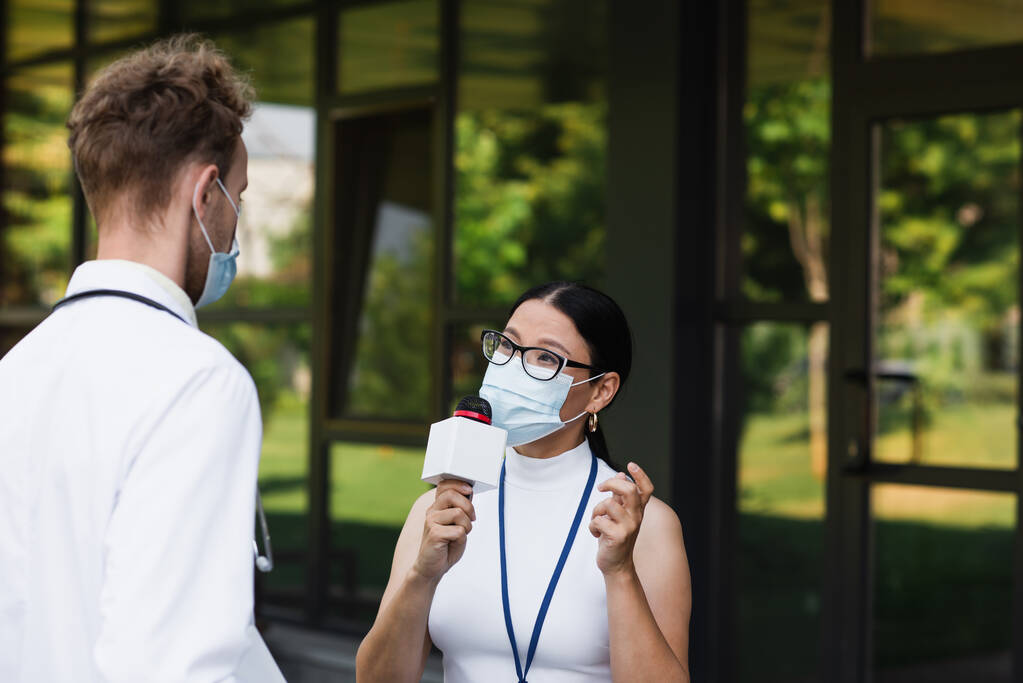 Tıbbi maskeli Asyalı muhabir, kliniğin yanındaki beyaz önlüklü doktorun yanında mikrofonla konuşuyor. - Fotoğraf, Görsel
