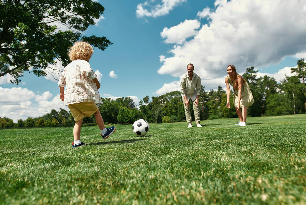 Νέοι γονείς διδάσκουν το μικρό τους γιο παίζοντας ποδόσφαιρο στο γρασίδι στο πάρκο μια καλοκαιρινή μέρα - Φωτογραφία, εικόνα