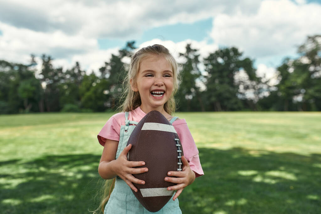 Portret wesołej dziewczynki trzymającej owalną, brązową, skórzaną piłkę rugby i uśmiechniętą podczas zabawy z rodzicami w parku - Zdjęcie, obraz