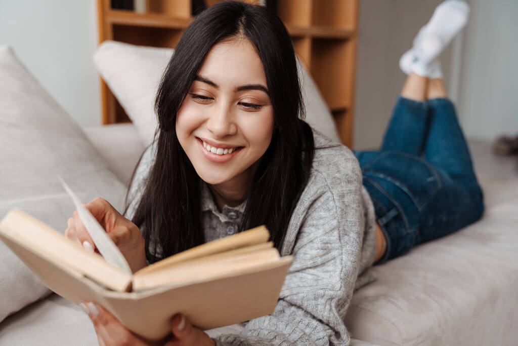 Fiatal boldog nő könyvet olvas, miközben ül a kanapén a nappaliban - Fotó, kép