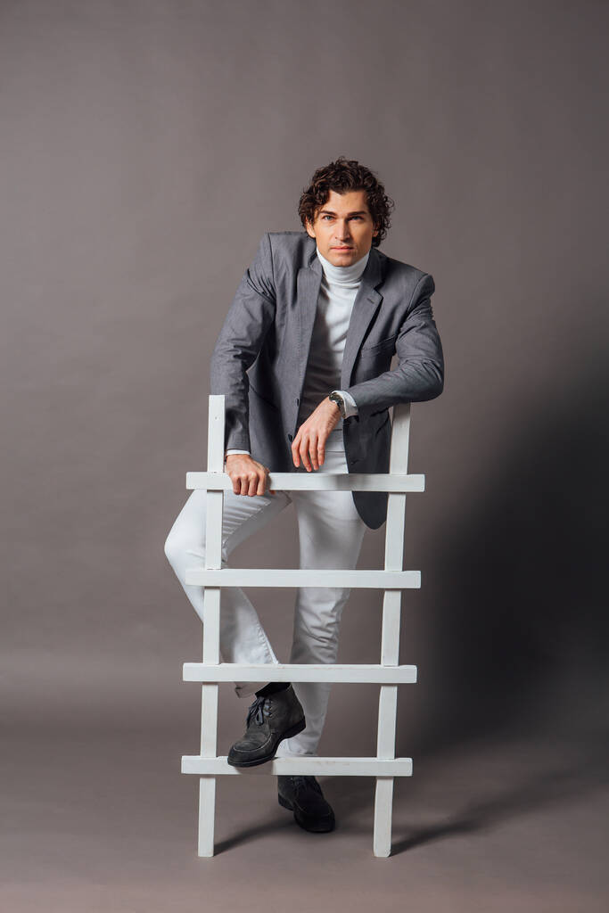 Fashion portrait of a tall handsome man dressed in white turtleneck, white jeans and grey jacket posing on the grey background with white ladder - Photo, Image