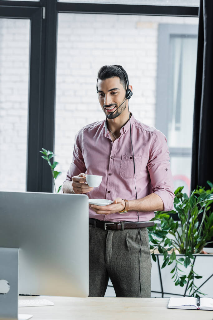 Pozitivní arabský podnikatel ve sluchátkách držící pohár během videohovoru na počítači v kanceláři  - Fotografie, Obrázek