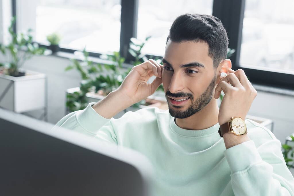Smiling arabian empresário segurando fone de ouvido sem fio perto de monitor de computador borrado no escritório  - Foto, Imagem