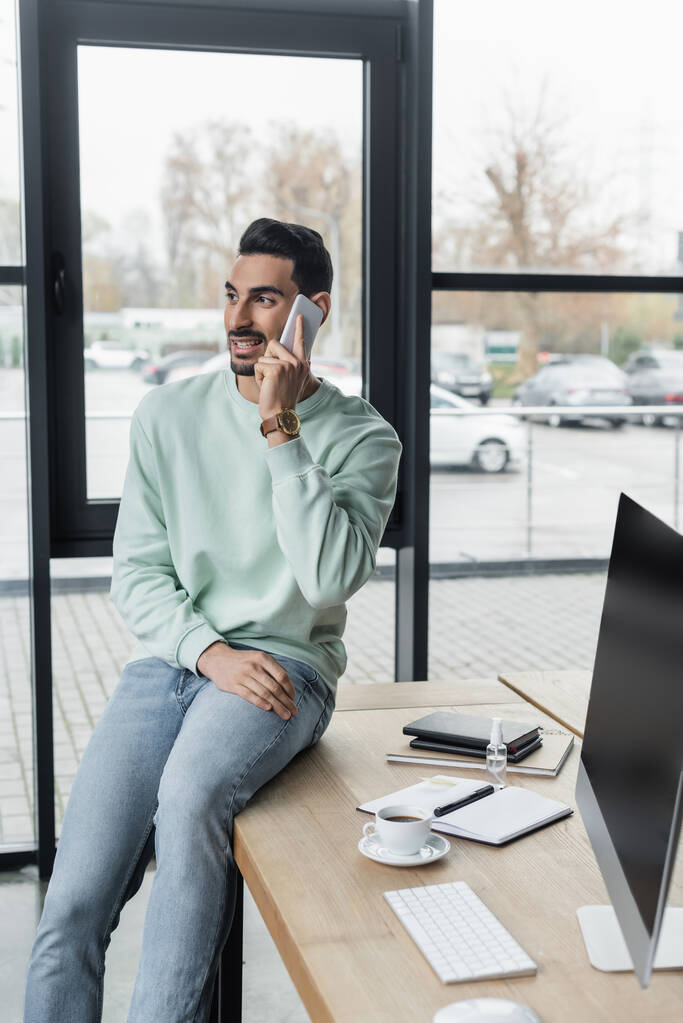 Hombre de negocios musulmán en ropa casual hablando en el teléfono inteligente cerca de portátiles y café en la oficina  - Foto, Imagen