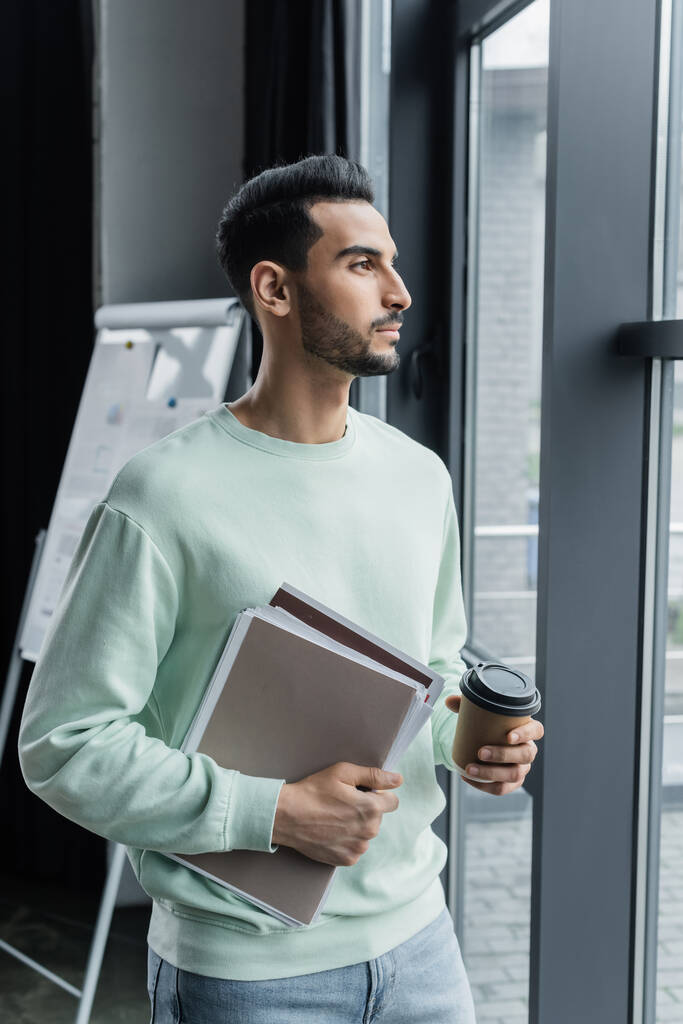 Young muslim manager in casual clothes holding paper folders and paper cup near window - Photo, Image