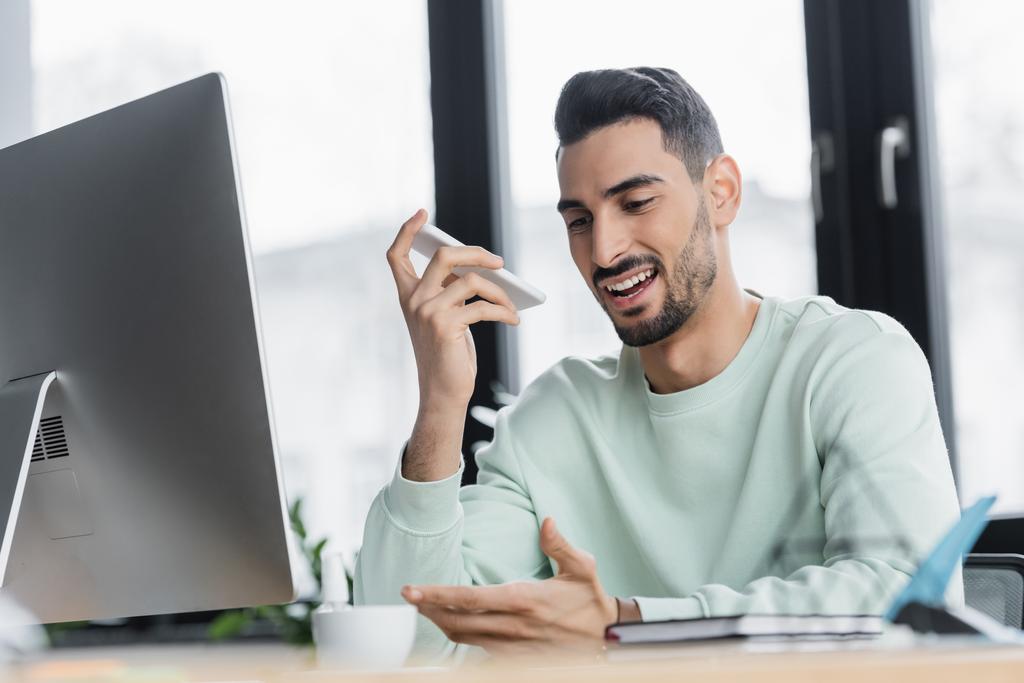 Uomo d'affari musulmano sorridente che registra il messaggio vocale sullo smartphone vicino al computer e al caffè in ufficio  - Foto, immagini