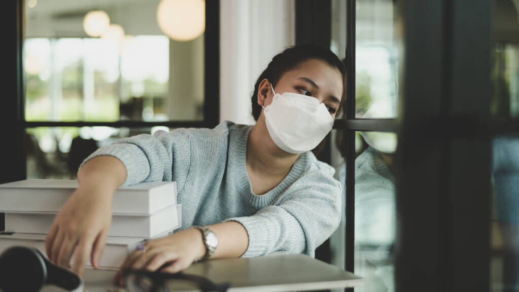 Eine asiatische Studentin mit medizinischer Maske sitzt am Fenster und schaut hinaus. - Foto, Bild