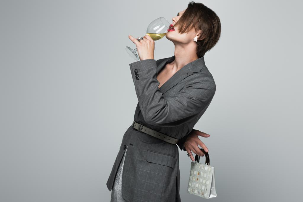 transznemű férfi blézerben bort iszik, miközben szürke bőröndöt tart - Fotó, kép