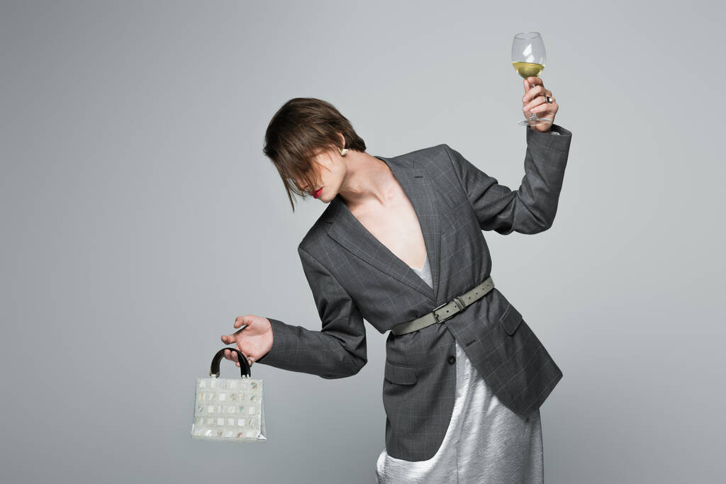 jeune homme transgenre en slip robe avec blazer tenant sac à main et verre de vin isolé sur gris - Photo, image