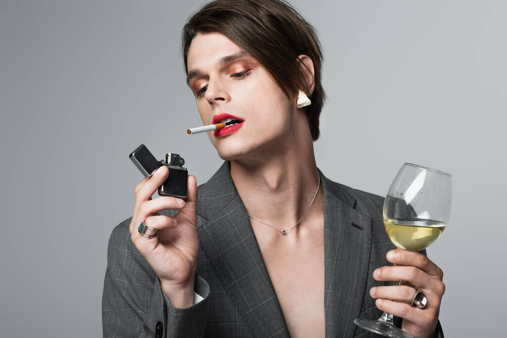 young transgender man in blazer holding glass of wine and lighter while smoking isolated on grey - Photo, Image