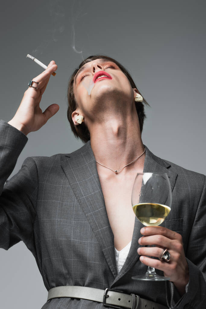 νεαρός τρανσέξουαλ άνδρας με σακάκι και σκουλαρίκια κρατώντας ένα ποτήρι κρασί ενώ το κάπνισμα απομονώνεται σε γκρι - Φωτογραφία, εικόνα