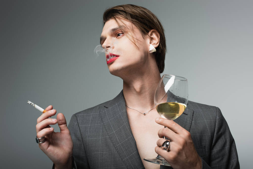 junger transsexueller Mann im Blazer hält Glas Wein und raucht Zigarette isoliert auf grau - Foto, Bild