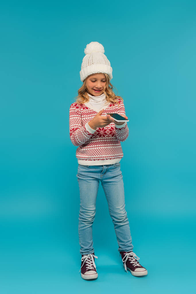 Ganzkörperansicht positiver Mädchen in warmem Pullover, die Sprachnachricht auf blau senden - Foto, Bild