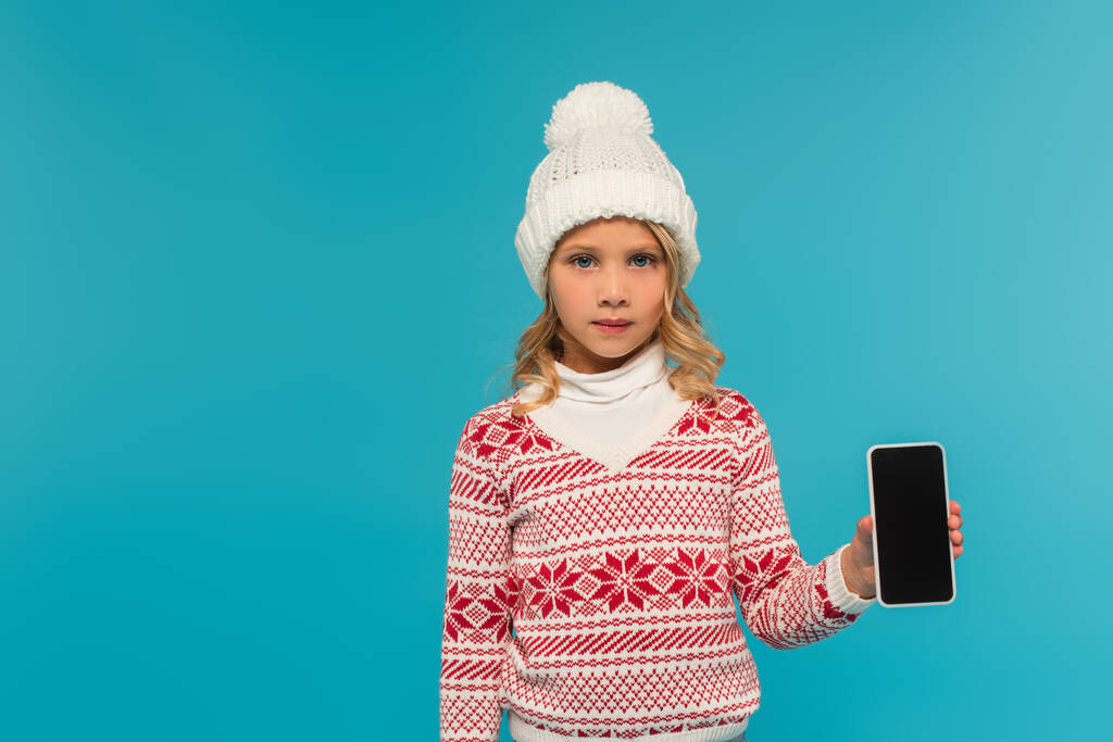 kid in knitted hat and sweater with ornament showing smartphone with blank screen isolated on blue - Photo, Image