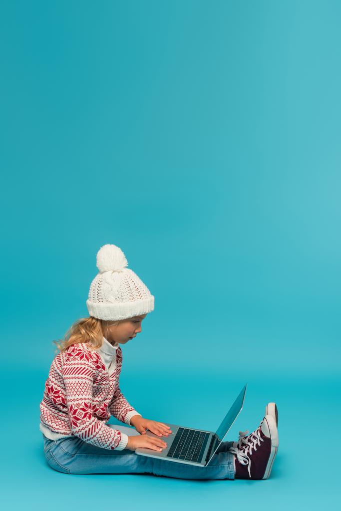 dziecko w ciepłym kapeluszu i swetrze za pomocą laptopa podczas siedzenia na niebiesko - Zdjęcie, obraz