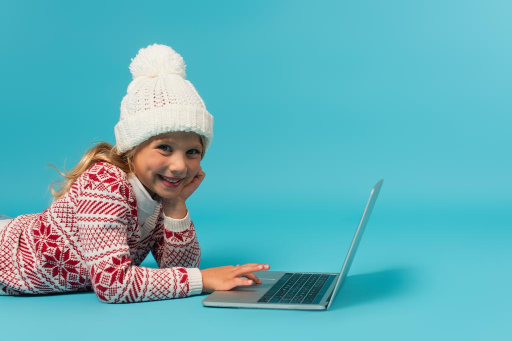 vrolijk meisje in gebreide hoed en trui met ornament met behulp van laptop terwijl liggend op blauw - Foto, afbeelding