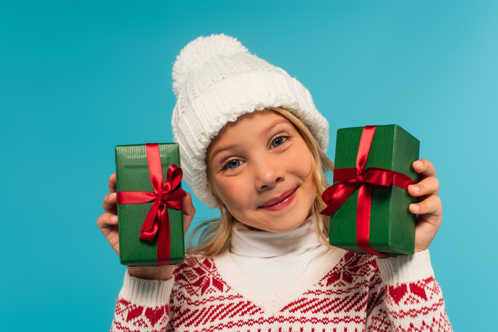 pleased girl in knitted hat holding green gift boxes isolated on blue - Photo, Image