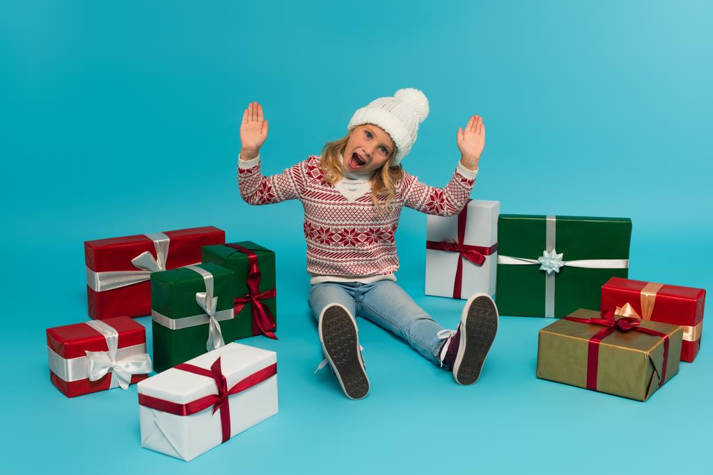Aufgeregtes Kind gestikuliert und blickt in die Kamera, während es neben bunten Geschenkboxen auf blauem Grund sitzt - Foto, Bild