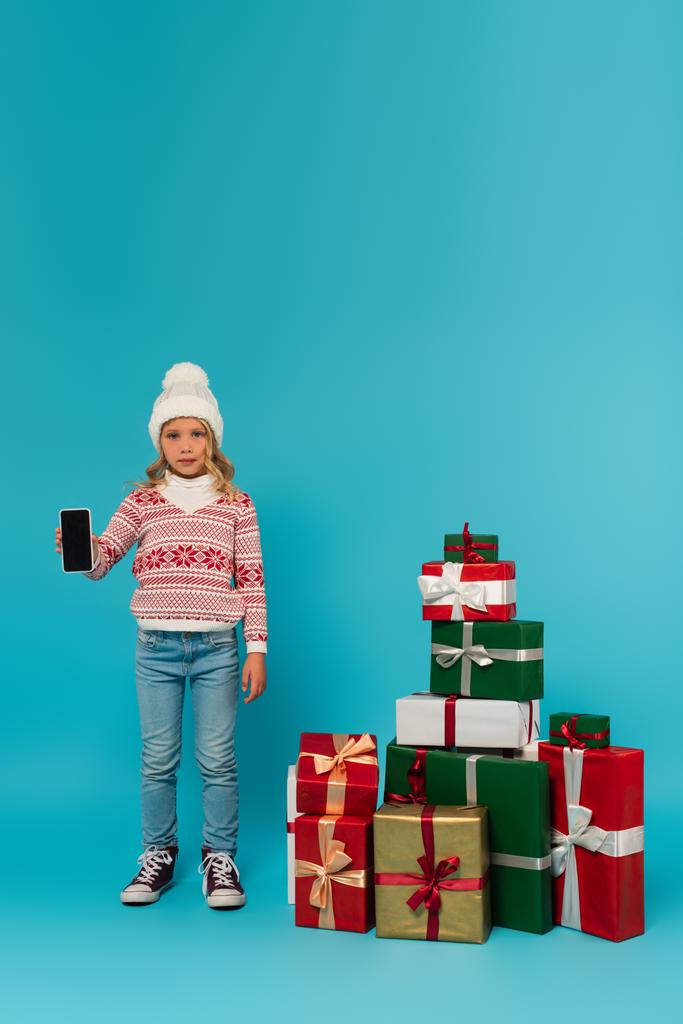 Sıcak kazaklı ve kot pantolonlu çocuk mavi ekranda boş ekranlı akıllı telefon gösteriyor. - Fotoğraf, Görsel