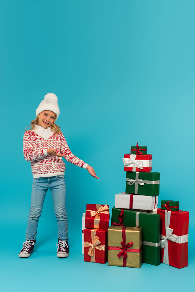 chica sonriente en sombrero de punto, suéter y jeans apuntando a la pila de regalos en azul - Foto, imagen