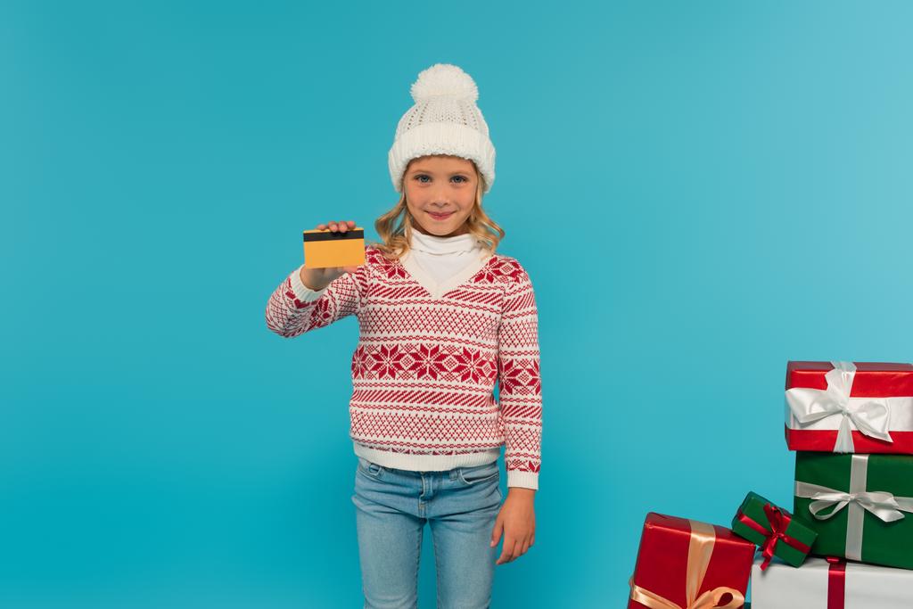 šťastná dívka v pletený klobouk a jumper držící kreditní kartu v blízkosti pestrobarevné dárkové krabice izolované na modré - Fotografie, Obrázek