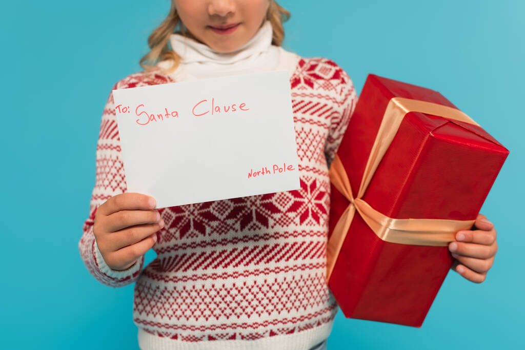 vista recortada de niño borroso con caja de regalo roja y carta a la cláusula de santa aislado en azul - Foto, imagen