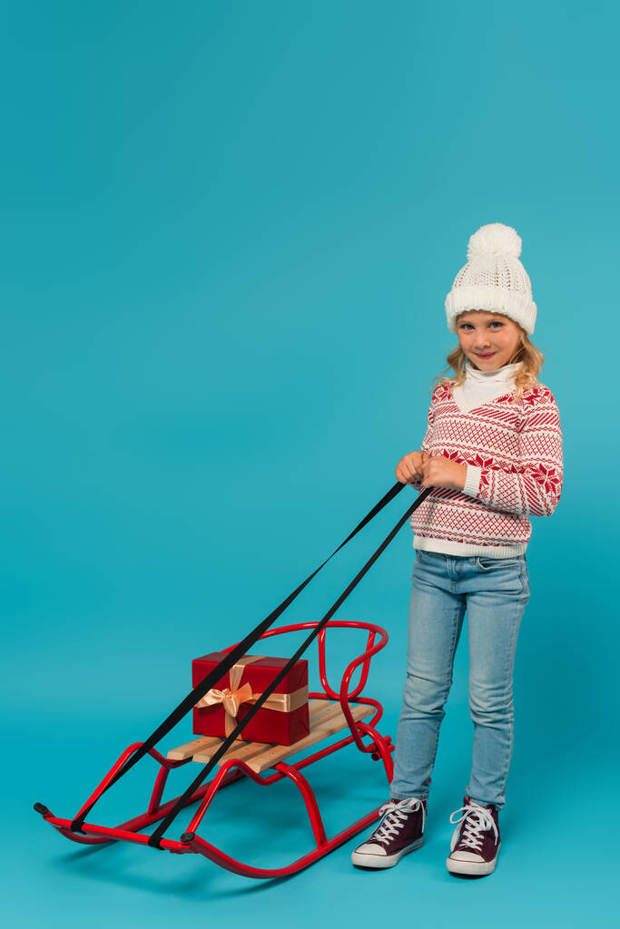 vista completa del niño en suéter caliente y jeans sonriendo cerca del trineo con caja de regalo en azul - Foto, imagen