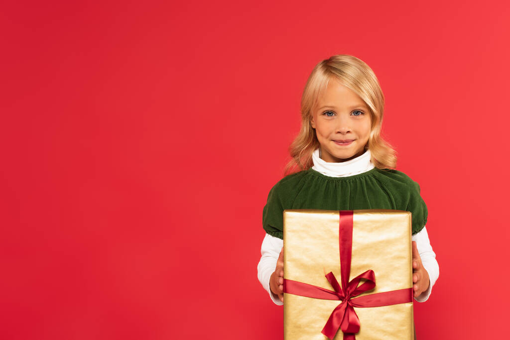 niño sonriente sosteniendo presente en papel de regalo dorado mientras mira a la cámara aislada en rojo - Foto, imagen