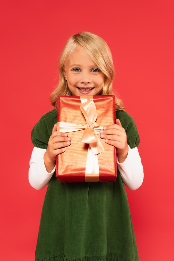 vrolijk meisje in groene jurk houden geschenk in gouden verpakking papier geïsoleerd op rood - Foto, afbeelding