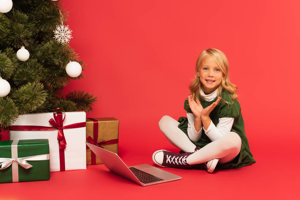 niño feliz aplaudiendo las manos cerca de la computadora portátil y regalos bajo el árbol de Navidad en rojo - Foto, imagen