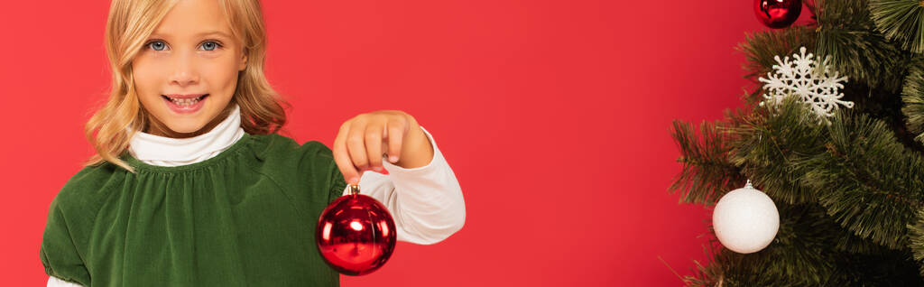uśmiechnięte dziecko trzymające błyszczącą świąteczną piłkę w pobliżu ozdobionego świerku odizolowanego na czerwono, sztandar - Zdjęcie, obraz