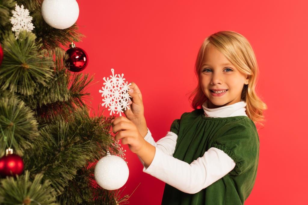 felice bambino tenendo fiocco di neve decorativo vicino albero di Natale isolato su rosso - Foto, immagini