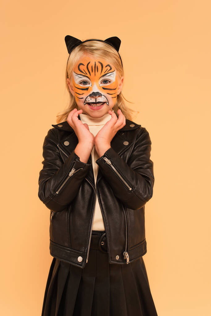 niño sonriente en chaqueta de cuero negro y maquillaje de tigre cogido de la mano cerca de la cara aislado en beige - Foto, Imagen