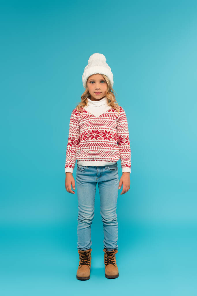 full length view of child in warm hat, jeans and sweater with ornament on blue - Photo, Image