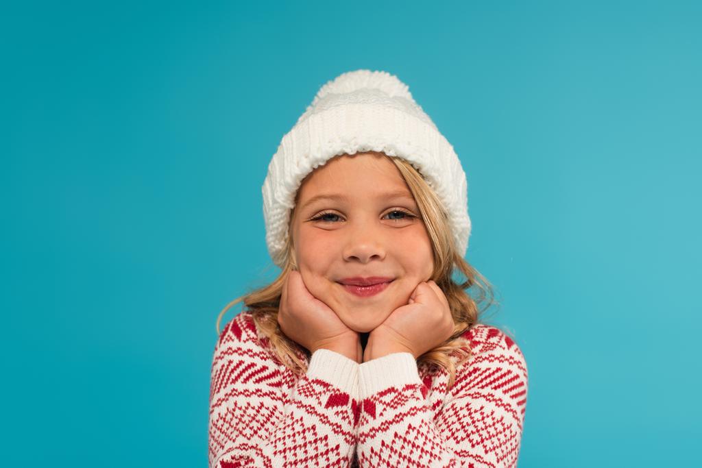 šťastný dítě v pleteném klobouku a teplý svetr s ozdobou při pohledu na kameru izolované na modré - Fotografie, Obrázek